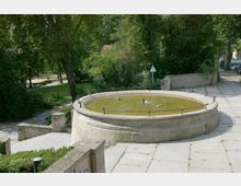 Runder Springbrunnen aus Stein mit gelblichem Wasser in einem parkähnlichen Bereich, umgeben von Bäumen und Gehwegen. Im Hintergrund sind Grünflächen, ein Fußweg und ein Verkehrszeichen sichtbar.