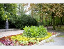 Ein gepflegter Park mit einem Blumenbeet aus gelben, roten und weißen Blumen im Vordergrund, flankiert von grünen Pflanzen. Im Hintergrund steht eine Büste auf einem Sockel, umgeben von dichten Büschen und Bäumen.