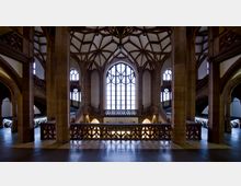 Historisches Innenraum einer gotischen Halle mit hohen Gewölbedecken, großen Fenstern mit filigranem Maßwerk und Bögen aus Sandstein. Eine zentrale Balustrade mit dekorativen Schnitzereien trennt verschiedene Ebenen, flankiert von geschwungenen Treppenaufgängen.