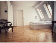 Helles Schlafzimmer mit minimalistischem Design, ausgestattet mit einem Bett, einem Holzstuhl und einem Einbauschrank. Der Raum hat eine schräge Decke mit zwei Dachfenstern, die viel Tageslicht auf den Holzboden werfen.