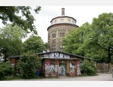 Ein historischer Wasserturm aus Backstein mit mehreren Etagen und einem schmalen Schornstein, umgeben von Bäumen. Im Vordergrund steht ein kleines, heruntergekommenes Gebäude mit Graffiti und Vegetation.