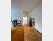 Ein moderner Innenraum mit hellem Fischgräten-Parkettboden, einem goldgelben Teppich, einem offenen Regal voller Bücher und Ordner sowie einem großen Wandspiegel. Entlang einer weißen Wand befinden sich Einbauschränke, und eine Tür im Hintergrund führt zu einem weiteren Bereich.