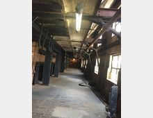 Industriegebäude mit einem langen Gang aus Ziegelmauern und Stahlträgern, beleuchtet von einer Deckenleuchte. An den Seiten verlaufen Rohre und Kabel, während rechts teilweise Fenster mit Blick nach draußen zu sehen sind.