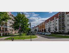 Wohnsiedlung mit mehrstöckigen Wohngebäuden in heller Fassade und Balkonen, umgeben von grünen Rasenflächen und Bäumen. Im Vordergrund eine gepflasterte Straße mit parkenden Autos und einem Wohnmobil.