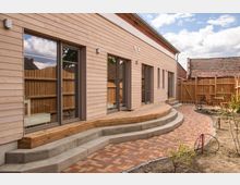 Moderne Holzfassade eines Gebäudes mit großen Fenstern und Stufen, die zu einer gepflasterten Terrasse führen. Der Bereich ist von einem Holzzaun umgeben und es sind einzelne Gartenpflanzen und Gartenmöbel zu sehen.