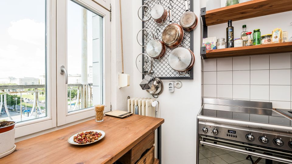 Helle Küche mit großer Fensterfront und Holzarbeitsplatte; an der Wand hängt ein Metallgitter mit Pfannen, daneben offene Holzregale mit Gewürzen und Gläsern. Rechts steht ein Edelstahlherd vor weiß gefliester Rückwand, unter dem Fenster sind Heizkörper und Balkonblick nach draußen zu sehen.
