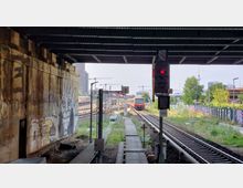 Blick auf eine Bahngleisanlage unter einer Brücke, begleitet von einer Signalampel mit rotem Licht im Vordergrund. Links eine mit Graffiti bedeckte Betonwand, rechts Zäune und Vegetation, im Hintergrund Gebäude und Baukräne.