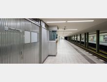 U-Bahn-Station mit zwei Bahnsteigen, grauem Fliesenboden und Säulen, die das flache Deckengewölbe stützen. Links im Bild befindet sich ein Dienstraum mit geriffelter Metallverkleidung und einer Tür.