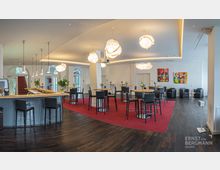 Moderner Veranstaltungsraum mit eleganter Bar und Sitzbereichen. Der Raum ist mit dunklem Holzboden, roten Teppichakzenten, dekorativen Hängelampen, runden Stehtischen sowie bunten Kunstwerken an den Wänden gestaltet.