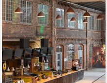 Innenansicht eines historischen Backsteingebäudes mit hohen Fenstern und industriellem Charme. Im Vordergrund befindet sich ein moderner Verkaufsbereich mit Bildschirmen und Hängelampen, während im Hintergrund eine Wand mit gemauerten Rundbögen und Dekorationen sichtbar ist.