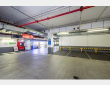 Untergeschoss eines Parkhauses mit markierten Parkplätzen, einer Ausfahrtbeschilderung, einem Parkautomaten sowie sichtbaren Lüftungsrohren und Leitungen an der Decke. Im Hintergrund Hinweise auf einen Supermarkteingang.