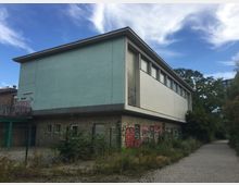 Ein zweistöckiges Gebäude mit Flachdach, einer weißen und türkisfarbenen Fassade sowie großen Glasbausteinen. Der untere Bereich ist mit Graffiti bedeckt, und das Gebäude steht in einer Umgebung mit Pflasterweg, Wildpflanzen und angrenzenden Bäumen.