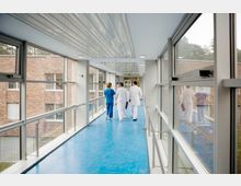 Ein moderner, gläserner Krankenhaus-Gang mit blauem Bodenbelag und großen Fenstern, die den Blick auf eine Backsteinfassade und Bäume freigeben. Drei medizinische Fachkräfte in weißer und blauer Kleidung gehen durch den Gang.