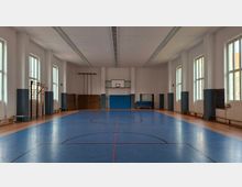 Leere Turnhalle mit blauem Sportboden und Markierungen, an der Stirnwand ein Basketballkorb mit Prallschutz. Hohe Fenster entlang der Seitenwände lassen Tageslicht herein; an den Wänden stehen Turngeräte, Matten und Bänke.