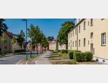 Wohnsiedlung mit gelb verputzten zweigeschossigen Reihenhäusern entlang einer Straße mit Fuß- und Radweg, flankiert von jungen Bäumen und gepflegten Grünflächen. Im Hintergrund sind weitere Häuser in Rosa zu erkennen, bei sonnigem Wetter unter blauem Himmel.