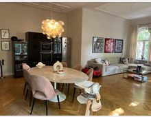 Offener Wohnbereich mit einem Esstisch aus hellem Marmor, umgeben von rosafarbenen Stühlen. Im Hintergrund eine moderne Küche mit schwarzen Schränken, eine große helle Couch, dekorative Bilder an der Wand und helle Fenster mit Blick ins Grüne.