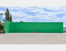 Flaches, offenes Gelände mit einer großen, grünen Wand im Hintergrund, die eine glatte Oberfläche hat und im Freien steht; davor ein kleines, rotes Auto auf einer betonierten Fläche; im Hintergrund blauer Himmel mit Wolken und einige Bäume.