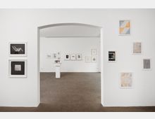 Weißer Galerieraum mit minimalistischer Einrichtung, in dem an den Wänden verschiedene gerahmte Kunstwerke hängen. Im Hintergrund steht ein kleiner weißer Sockel mit darauf platzierten Objekten, brauner Boden und ein weißer Durchgangsbereich sind sichtbar.