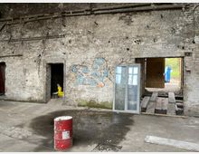 Verlassene Lagerhalle mit nackten, verwitterten Ziegelwänden und sichtbaren Rohren. In der Halle befinden sich eine rote Metalltonne, ein verschmutzter Boden mit Wasserpfütze, sowie offene Türöffnungen, teilweise mit Graffiti und freigelegtem Außenbereich.