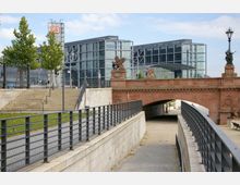 Moderne Glasfassade des Berliner Hauptbahnhofs im Hintergrund, davor eine historische Brücke aus rotem Sandstein mit dekorativen Statuen. Im Vordergrund Treppen und Wege mit Geländern, die unter der Brücke entlangführen.