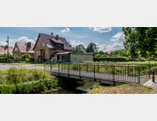 Ein kleines Wohnhaus steht in einem ländlichen Gebiet mit Vorgarten und einer angrenzenden Wiese. Im Vordergrund führt eine Metallbrücke über einen schmalen Bach, umgeben von hohen Bäumen und Sträuchern.