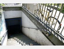 Abgang zu einer Unterführung mit Stufen, weißen Kacheln an den Wänden und einem blauen Gittertor am unteren Ende. Oberhalb des Abgangs befinden sich ein Geländer und ein gelbes Fahrrad.