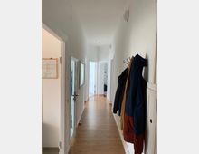 Ein heller Flur mit weiß gestrichenen Wänden und Holzboden, gesäumt von Türen zu anderen Räumen. Rechts hängt eine Garderobe mit mehreren Jacken, links ist ein Spiegel und ein teilweise geöffneter Raum sichtbar.