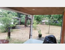 Blick von einer überdachten Terrasse auf einen Garten mit trockener Wiese, Bäumen und Sträuchern. Im Vordergrund steht eine alte Handpumpe, im Hintergrund sind ein Baumhaus mit Rutsche, kleine Gebäude und ein geparktes Fahrzeug zu sehen.