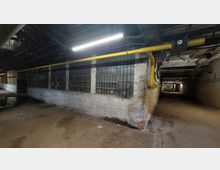 Unterirdischer Gang mit groben Betonwänden, einem vergitterten Fensterbereich aus Glasbausteinen und einer gelben Leitung, die entlang der Decke verläuft. Der Raum ist schwach beleuchtet und zeigt sichtbare Abnutzungsspuren.