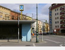 Straßenszene mit einem U-Bahn-Zugangsgebäude der Linie U7, erkennbar an einem blauen Ziegelmauerwerk und einem gelben U-Bahn-Schild. Im Hintergrund mehrstöckige Wohnhäuser mit Balkonen und Geschäften in einem urbanen Viertel.
