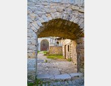 Steinbogen einer historischen Burgmauer, unter dem eine gepflasterte Fläche und ein Weg zu weiteren Burggebäuden führen. Im Hintergrund sind ein weiterer Rundbogen, eine Holztür und eine Wiese sichtbar, umgeben von alten Steinmauern.