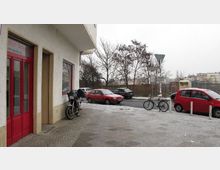 Eine Straßenansicht im Winter mit einem Bürgersteig, auf dem ein Fahrrad und ein Motorrad abgestellt sind, sowie einem Gebäude mit roten Türen links im Bild. Im Hintergrund sind geparkte Autos, schneebedeckter Boden und kahle Bäume zu sehen.