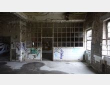 Ein verlassener Raum mit bröckelnden Wänden, großen Metallgittern vor Fenstern und weißen Fliesen, die mit Graffiti besprüht sind. Der Boden ist verschmutzt, und die Umgebung wirkt heruntergekommen und stark verfallen.