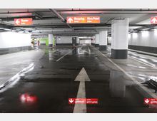 Unterirdisches Parkhaus mit beleuchteten roten Schildern, die zu „Shopping“, „Kino/Hotel“ und „Theater“ weisen. Der Boden ist nass und spiegelt das Licht, die weiße Fahrbahnmarkierung zeigt geradeaus.