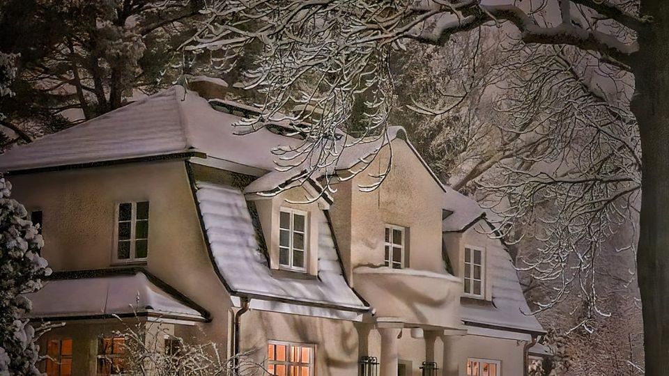 Verschneites zweistöckiges Wohnhaus mit steilem Satteldach und mehreren Gaubenfenstern, vor einem Zaun und winterlichen Sträuchern. Große, schneebedeckte Baumäste rahmen die Szene ein, die Fenster leuchten warm in einer nächtlichen Umgebung.