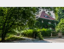 Ein zweistöckiges Wohnhaus mit rotem Satteldach, dessen Fassade dicht mit Efeu bewachsen ist, umgeben von dichtem Grün aus Büschen und Bäumen. Im Vordergrund verlaufen ein Gehweg und eine Straße unter schattigen Baumkronen.