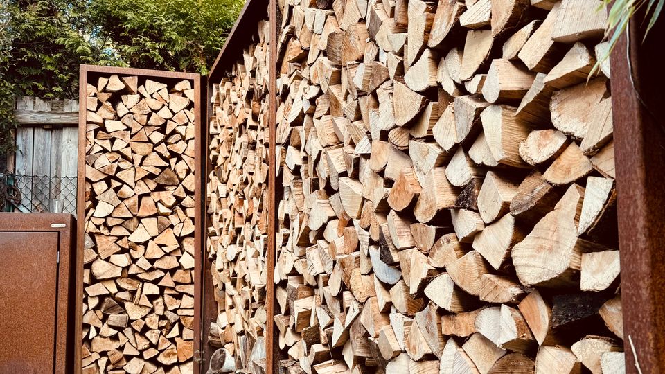 Schmaler Außenbereich mit einem langen Holzlager, in dem gespaltene Scheite dicht in einem Metallrahmen gestapelt sind, sowie einem passenden Holzstapel-Element am Ende. Gepflasterter Boden, Zaun und Bäume im Hintergrund unter blauem Himmel.