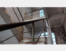 Innenansicht eines verfallenen Gebäudes mit einer alten, rostigen Treppe aus Metall und Beton. Die Wände zeigen abbröckelnden Putz und dahinterliegende Mauersteine, während durch große Fenster Tageslicht einfällt.