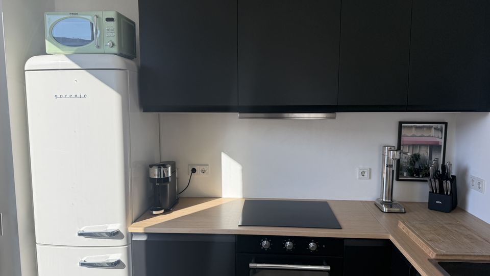 Moderne Küchenzeile mit mattschwarzen Hängeschränken, Holzarbeitsplatte und schwarzem Kochfeld über einem Einbauofen. Links steht ein weißer Retro-Kühlschrank mit Mikrowelle, auf der Arbeitsfläche befinden sich Kaffeemaschine, Wasserhahn/Spender, Messerblock und ein gerahmtes Bild vor einer weißen Wand mit Steckdosen.