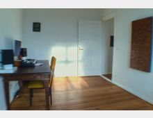 Helles Arbeitszimmer mit Holzdielenboden, einem schlichten Holztisch mit gelber Stuhlpolsterung und einem Monitor darauf; im Hintergrund eine weiße Tür, ein Spiegel und ein hölzernes Wanddekor.