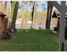 Ein Garten mit einer Rasenfläche, umgeben von hohen Kiefern, und Blick auf einen See im Hintergrund. Auf der linken Seite ist ein Gebäude mit Holzverkleidung, rechts eine überdachte Terrasse, dahinter sind Bäume und eine Uferpromenade erkennbar.