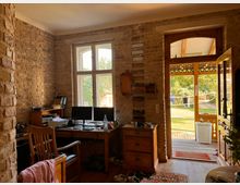 Ein rustikales Zimmer mit Wänden aus sichtbaren Backsteinen. Links steht ein Holzschreibtisch mit zwei Monitoren und Büromaterial, daneben eine Holzkommode. Rechts führt eine geöffnete Tür mit Glaseinsatz auf eine Veranda, dahinter sieht man einen Garten mit Bäumen und einem Volleyballnetz.