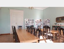 Ein heller Raum mit hohen Decken, ausgestattet mit langen Holztischen und verzierten Stühlen. An der mintgrünen Wand hängen Infotafeln mit Fotos und Texten; eine weiße Tür und ein Kamin mit Dekorationen sind ebenfalls erkennbar.