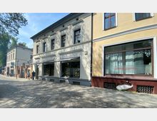 Straßenansicht mit Kopfsteinpflaster, flankiert von zweistöckigen historischen Gebäuden. Links befindet sich ein Geschäft mit großen Schaufenstern, während rechts ein Fenster mit Werbeaufschrift für Kosmetikleistungen sichtbar ist; im Hintergrund sind Bäume und ein weiterer Gebäudeeingang zu sehen.