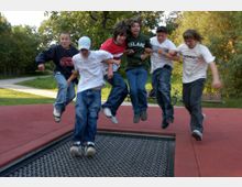 Eine Gruppe von sechs Jugendlichen springt gemeinsam auf einem im Boden eingelassenen Trampolin in einem Park. Im Hintergrund sind Bäume, Wiesen, ein Weg und eine Parkbank zu sehen.