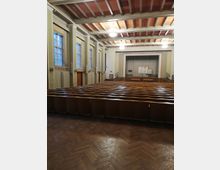 Ein Vortragssaal mit einer Bühne im Vordergrund, Sitzreihen aus Holz und hohen Fenstern an den Seiten. Die Decke ist mit sichtbaren Balken und dezenten Leuchtern gestaltet, der Boden besteht aus Holzparkett.