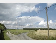 Ländliche Szenerie mit einer unbefestigten Straße, die sich an einer Weggabelung durch eine offene Landschaft erstreckt. Holzmasten mit Stromleitungen ziehen sich entlang der Straße, flankiert von Wiesen, Feldern und vereinzelten Gebäuden im Hintergrund.