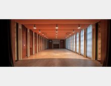 Langrechteckiger Saal mit Holzparkettboden und einer hohen Fensterreihe mit Vorhängen auf der rechten Seite, links von vertikal gegliederten Holzpaneelen gesäumt; die Decke ist orange gestrichen und mit mehreren Hängeleuchten ausgestattet. Im Hintergrund befindet sich eine Holzverkleidung mit Türen und einem kleinen Fensterausschnitt.
