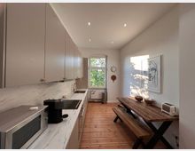 Moderne Küche mit hellen Wänden und Holzfußboden. Links befinden sich beige Einbauschränke und eine Arbeitsplatte in Marmoroptik mit Küchengeräten, rechts ein Tisch und eine Bank aus Holz, darüber hängt eine Zeichnung an der Wand. Ein Fenster im Hintergrund lässt Tageslicht herein.