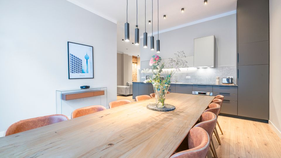 Heller, modern eingerichteter Ess- und Küchenbereich mit langem Holztisch und gepolsterten Stühlen, darüber mehrere zylindrische Pendelleuchten. Im Hintergrund eine minimalistische Einbauküche mit grauen Fronten, hoher Schrankwand, Arbeitsflächenbeleuchtung und Holzfußboden.